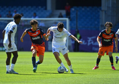 Soccer players in white and orange kits contesting the ball on a sunny pitch near the goal.