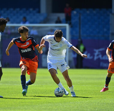 Soccer players in white and orange kits contesting the ball on a sunny pitch near the goal.