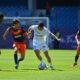 Soccer players in white and orange kits contesting the ball on a sunny pitch near the goal.