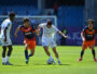 Soccer players in white and orange kits contesting the ball on a sunny pitch near the goal.