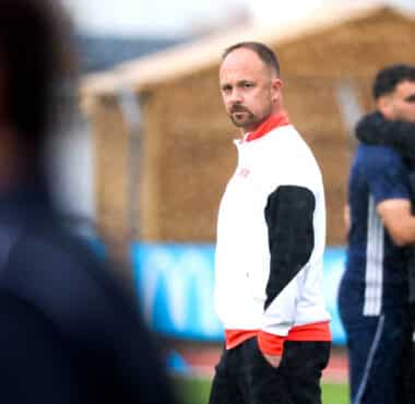 Man in a white and red athletic jacket stands on a field, looking toward the camera as blurred teammates hug in the background.