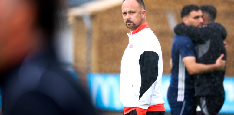Man in a white and red athletic jacket stands on a field, looking toward the camera as blurred teammates hug in the background.