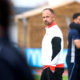 Man in a white and red athletic jacket stands on a field, looking toward the camera as blurred teammates hug in the background.