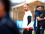 Man in a white and red athletic jacket stands on a field, looking toward the camera as blurred teammates hug in the background.