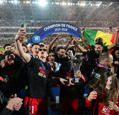 Celebrating football team holding a trophy beneath a blue 'Champion de France 2025-2026' arch amid confetti.