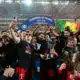 Celebrating football team holding a trophy beneath a blue 'Champion de France 2025-2026' arch amid confetti.