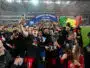 Celebrating football team holding a trophy beneath a blue 'Champion de France 2025-2026' arch amid confetti.