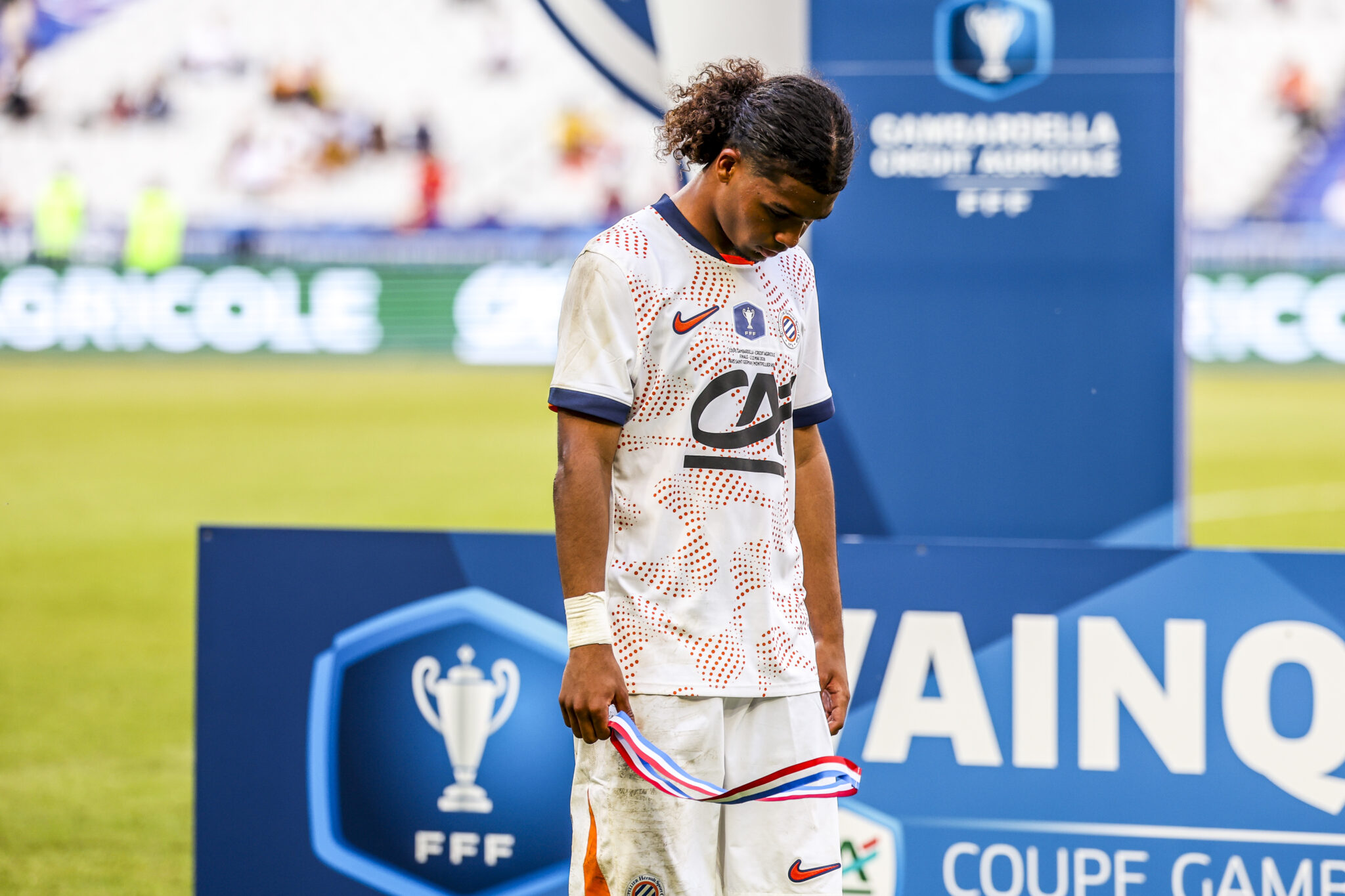 Paris Saint-Germain v Montpellier HSC - Gambardella Credit Agricole Cup Final 2026 | AllezPaillade.com Young soccer player in white jersey with orange dots on a podium, holding a striped ribbon after a trophy ceremony.