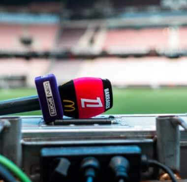 Close-up of a red and purple sports microphone resting on a control desk with blurred stadium seating in the background.