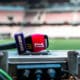 Close-up of a red and purple sports microphone resting on a control desk with blurred stadium seating in the background.