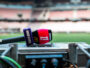 Close-up of a red and purple sports microphone resting on a control desk with blurred stadium seating in the background.