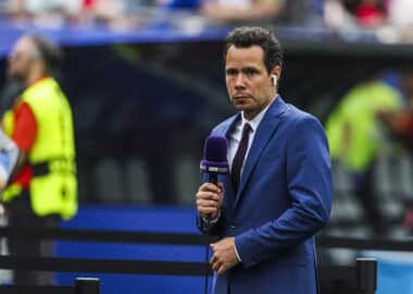 Male sports presenter in a blue suit with a purple microphone and wireless earbud, reporting on the field with stadium crowds behind him.