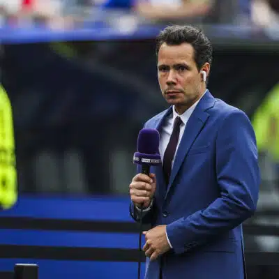 Male sports presenter in a blue suit with a purple microphone and wireless earbud, reporting on the field with stadium crowds behind him.
