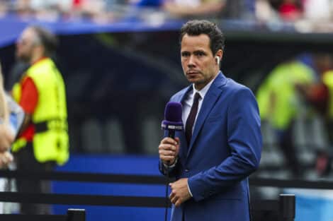 Male sports presenter in a blue suit with a purple microphone and wireless earbud, reporting on the field with stadium crowds behind him.