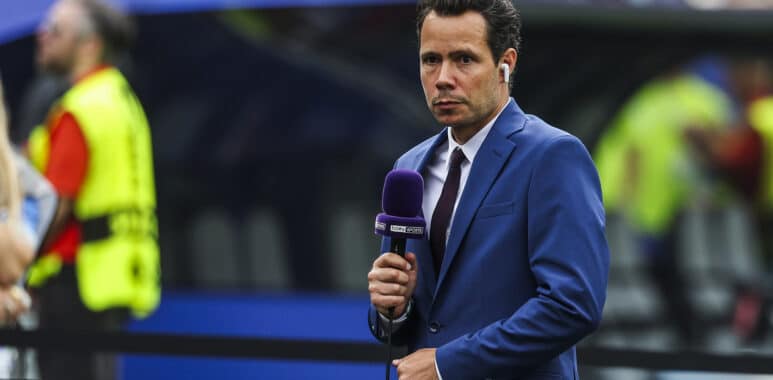 Male sports presenter in a blue suit with a purple microphone and wireless earbud, reporting on the field with stadium crowds behind him.