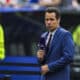 Male sports presenter in a blue suit with a purple microphone and wireless earbud, reporting on the field with stadium crowds behind him.