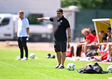 Soccer coach on the sideline pointing, with players on the bench and multiple balls on the grass.