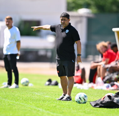 Soccer coach on the sideline pointing, with players on the bench and multiple balls on the grass.
