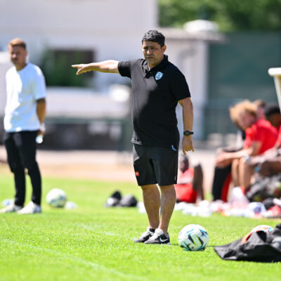 Soccer coach on the sideline pointing, with players on the bench and multiple balls on the grass.