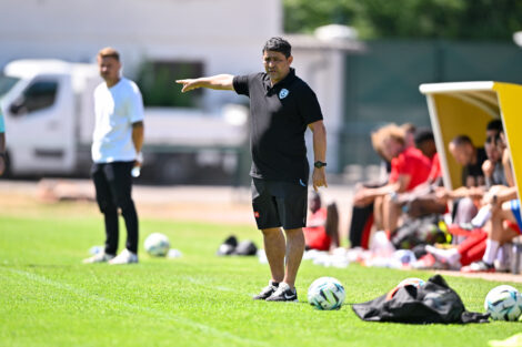 Soccer coach on the sideline pointing, with players on the bench and multiple balls on the grass.