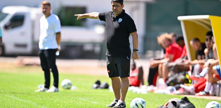 Soccer coach on the sideline pointing, with players on the bench and multiple balls on the grass.