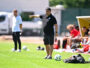 Soccer coach on the sideline pointing, with players on the bench and multiple balls on the grass.
