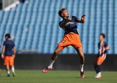 Soccer player leaping mid-air during training, wearing a navy jersey and bright orange shorts on a green field with blue stadium seating behind.