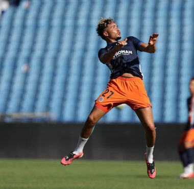 Soccer player leaping mid-air during training, wearing a navy jersey and bright orange shorts on a green field with blue stadium seating behind.