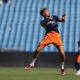 Soccer player leaping mid-air during training, wearing a navy jersey and bright orange shorts on a green field with blue stadium seating behind.