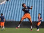 Soccer player leaping mid-air during training, wearing a navy jersey and bright orange shorts on a green field with blue stadium seating behind.