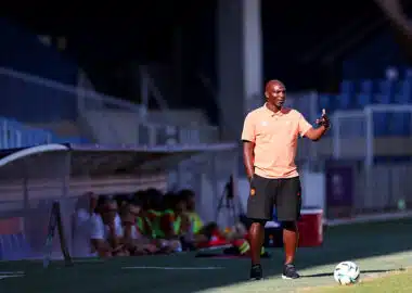 Soccer coach on the sideline gesturing with one hand, wearing a peach polo and black shorts, with a ball at his feet on a sunny field; players sit on a bench blurred in the background.