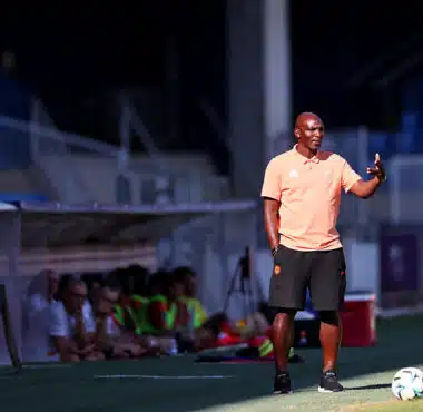 Soccer coach on the sideline gesturing with one hand, wearing a peach polo and black shorts, with a ball at his feet on a sunny field; players sit on a bench blurred in the background.