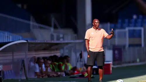Soccer coach on the sideline gesturing with one hand, wearing a peach polo and black shorts, with a ball at his feet on a sunny field; players sit on a bench blurred in the background.