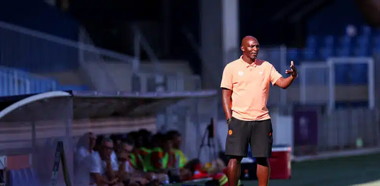 Soccer coach on the sideline gesturing with one hand, wearing a peach polo and black shorts, with a ball at his feet on a sunny field; players sit on a bench blurred in the background.