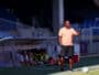 Soccer coach on the sideline gesturing with one hand, wearing a peach polo and black shorts, with a ball at his feet on a sunny field; players sit on a bench blurred in the background.