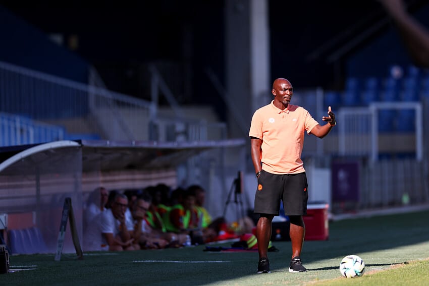 Soccer coach on the sideline gesturing with one hand, wearing a peach polo and black shorts, with a ball at his feet on a sunny field; players sit on a bench blurred in the background.