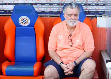 Older man with gray hair and beard sits in a dugout on orange-and-blue seats, wearing a coral polo and glasses hanging from his shirt.