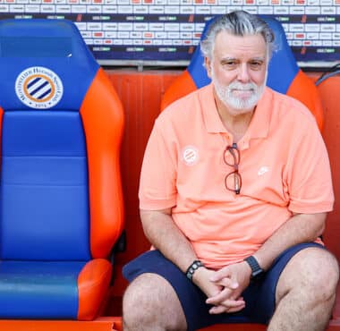 Older man with gray hair and beard sits in a dugout on orange-and-blue seats, wearing a coral polo and glasses hanging from his shirt.