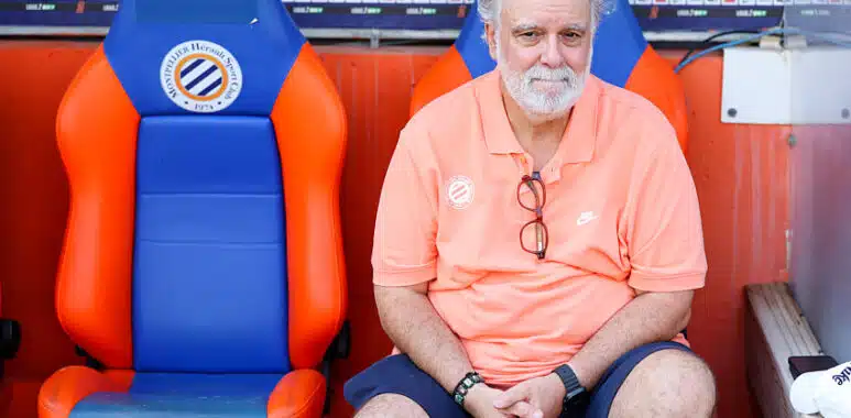 Nicollin | AllezPaillade.com Older man with gray hair and beard sits in a dugout on orange-and-blue seats, wearing a coral polo and glasses hanging from his shirt.