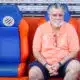 Older man with gray hair and beard sits in a dugout on orange-and-blue seats, wearing a coral polo and glasses hanging from his shirt.