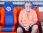 Older man with gray hair and beard sits in a dugout on orange-and-blue seats, wearing a coral polo and glasses hanging from his shirt.