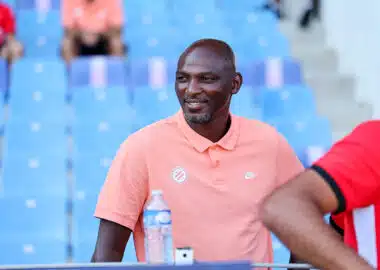 Man wearing a peach polo sitting in blue stadium seats, smiling, with a water bottle on the table nearby.