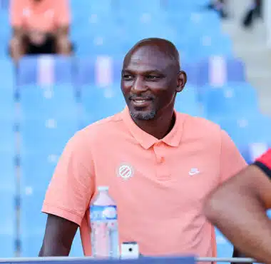 Man wearing a peach polo sitting in blue stadium seats, smiling, with a water bottle on the table nearby.