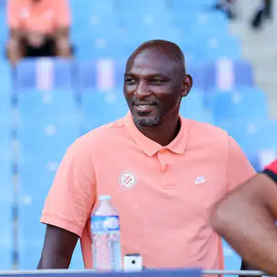 Man wearing a peach polo sitting in blue stadium seats, smiling, with a water bottle on the table nearby.