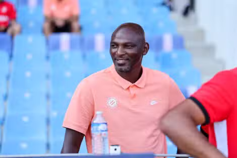 Man wearing a peach polo sitting in blue stadium seats, smiling, with a water bottle on the table nearby.