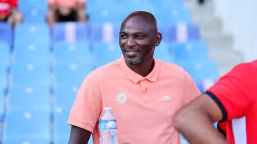 Man wearing a peach polo sitting in blue stadium seats, smiling, with a water bottle on the table nearby.