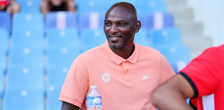 Camara | AllezPaillade.com Man wearing a peach polo sitting in blue stadium seats, smiling, with a water bottle on the table nearby.