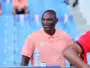 Man wearing a peach polo sitting in blue stadium seats, smiling, with a water bottle on the table nearby.
