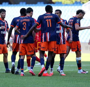 Soccer players in blue and orange jerseys form a huddle on the green field during a match. One player with number 11 stands central. Stadium seats are blurred in the background.