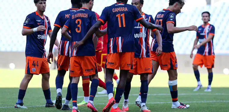 Soccer players in blue and orange jerseys form a huddle on the green field during a match. One player with number 11 stands central. Stadium seats are blurred in the background.